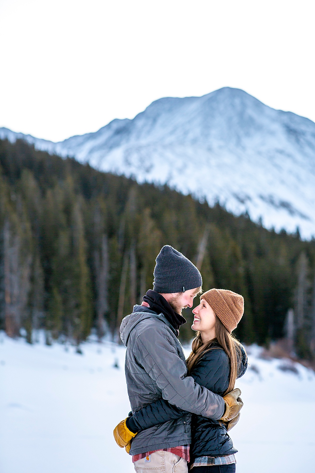 Couple hugs with snowy mountain in the background during a winter engagement shoot in Breckenridge, Colorado taken by Hillary Shedd Photography, Colorado Wedding Photographer.